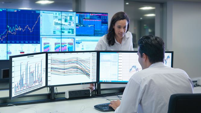 back of a man sitting at a desk with keyboard in front of 3 large monitors, with a standing woman facing him, in front of a bank of monitors